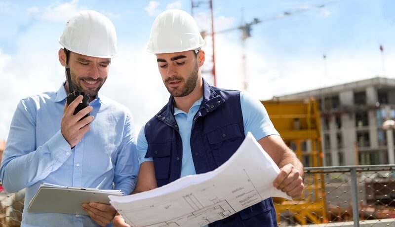 Zwei Bauleiter mit Helm prüfen Bauplan auf Baustelle, Funkgerät in Hand